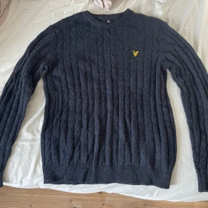 Mörkgrå stickad tröja från Lyle & Scott - Säljer en snygg mörkgrå stickad tröja från Lyle & Scott med kabelstickat mönster. Tröjan har en gul broderad logotyp på bröstet och långa ärmar. Perfekt för kyligare dagar! Köpte den i vintras och trodde jag skulle använda den men så blev de aldrig. Den är bara testad! Nypris 1199kr!