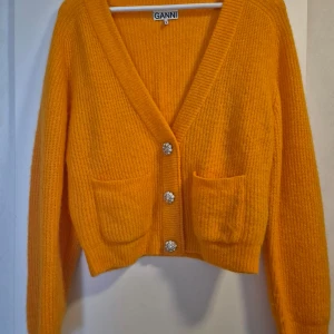 Orange ullstickad kofta från Ganni - Säljer en mjuk och stilren orange ullstickad kofta från Ganni. Koftan har en v-ringad design med dekorativa knappar och praktiska fickor framtill. Perfekt för att hålla sig varm och trendig.