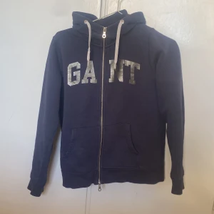 Mörkblå Gant hoodie   - Säljer en snygg mörkblå hoodie från GANT med dragkedja och stor logga på bröstet. Tröjan har långa ärmar och en praktisk huva med snören. Perfekt för en avslappnad stil.