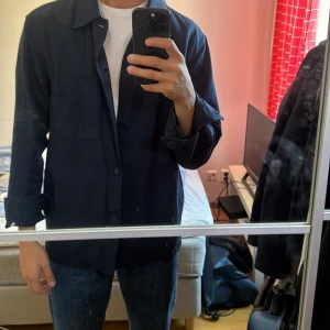 Mörkblå overshirt från Uniqlo - Säljer en stilren mörkblå overshirt från Uniqlo. Skjortan har långa ärmar och knappar framtill. Perfekt för lager-på-lager-stil och passar till många olika outfits. Använd 4 gånger och är perfekt nu till sommaren med skjorta eller med en vanlig t-shirt M storlek som sytts in som S