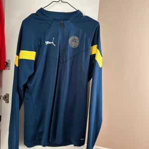 Mörkblå långärmad tröja från Puma - Säljer en mörkblå långärmad tröja från Puma med gula detaljer på ärmarna. Tröjan har en dragkedja vid halsen och ett tryckt emblem på bröstet. Perfekt för sportiga aktiviteter eller som en stilren vardagströja.