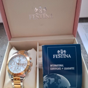 Festina klocka i guld och silver - Säljer en elegant Festina klocka med en kombination av guld och silver på armbandet. Urtavlan är vit med flera sub-dials och guldfärgade detaljer. Klockan kommer i originalförpackning med internationellt garantibevis.