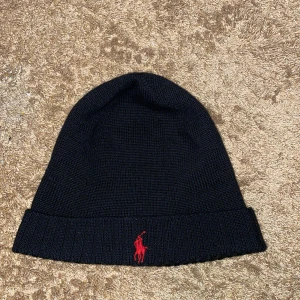 Svart mössa från Polo Ralph Lauren - Snygg svart mössa från Polo Ralph Lauren med röd broderad logga framtill. Tillverkad i mjukt material för en bekväm passform. Perfekt för kyliga dagar när det är vinter ❄️pris kan diskuteras vid snabba affär😃