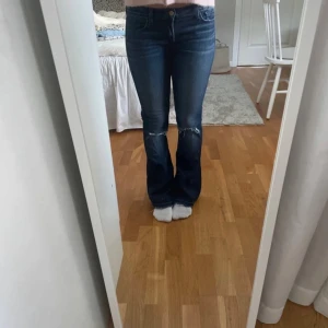 Blå bootcut jeans - Snygga blå bootcut jeans med låg midja och slitna detaljer vid knäna. Perfekta för en avslappnad stil. De har en klassisk femficksdesign och stängs med dragkedja och knapp.