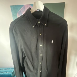 Ralph Lauren skjorta  - Snygg svart skjorta från Ralph Lauren med klassisk krage och knappar framtill. Skjortan har en broderad logotyp på bröstet och är långärmad. Perfekt för en stilren look i storlek M. Skick 9/10✅ Skriv vid frågor eller funderingar i DM🍾