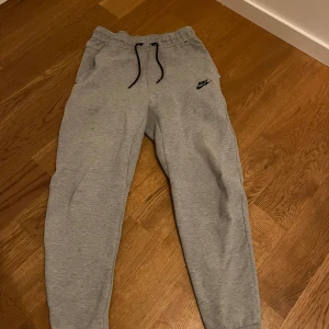 Grå Nike tech Byxor - Säljer ett par grå mjukisbyxor från Nike med svart logga på sidan. De är tillverkade i mjukt material för extra komfort. Hör av dig vid funderingar!