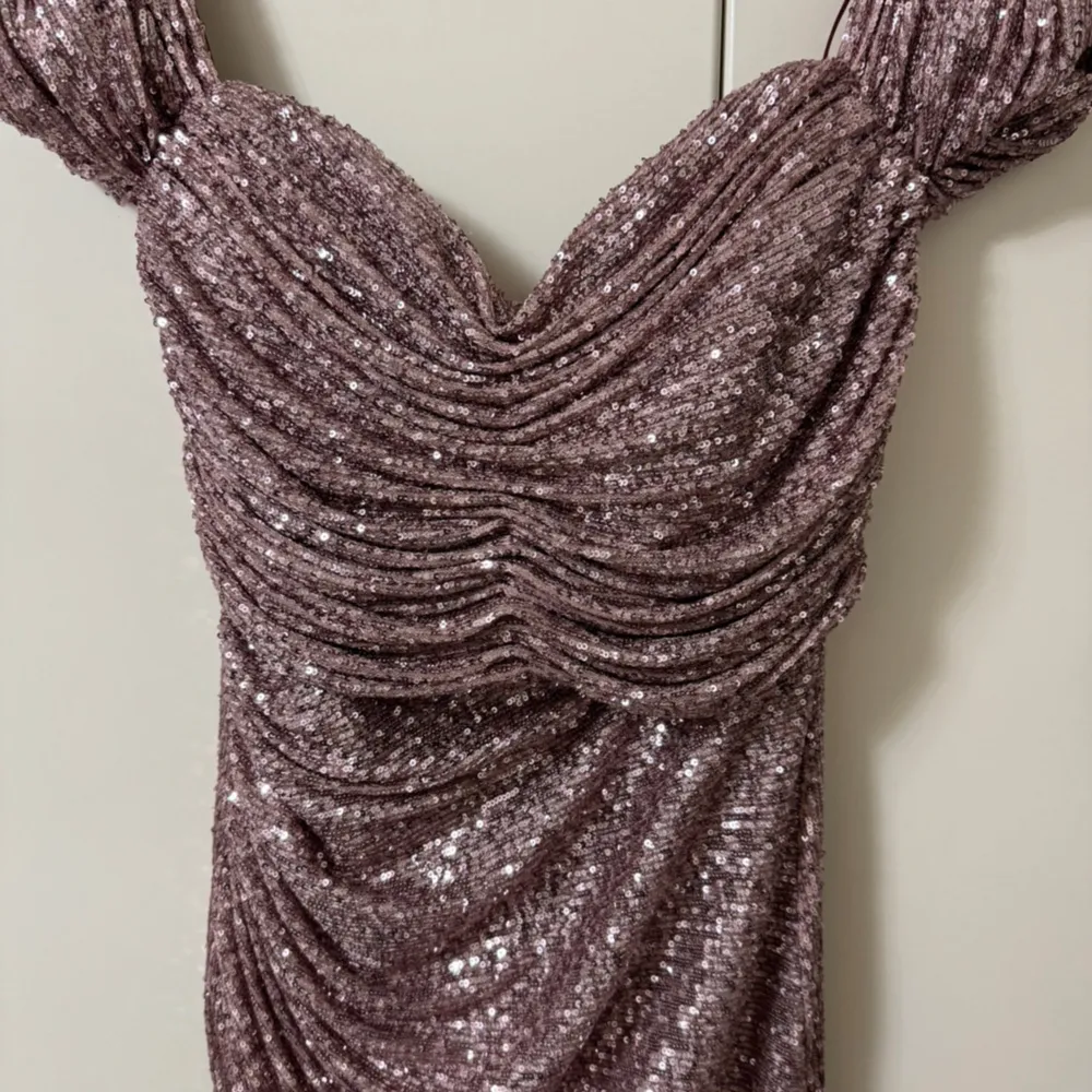 Säljer en fantastisk glittrig rosa långklänning med draperad design. Klänningen har en vacker omlott-look och är perfekt för festliga tillfällen. Den är axelbandslös och har en elegant passform som framhäver figuren.. Mekot.