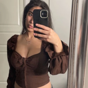 Brun långärmad topp - Snygg brun långärmad topp med knytning framtill och en croppad passform. Toppen har en djup v-ringning och voluminösa ärmar som ger en trendig look.