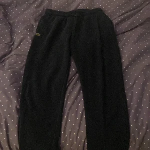 Mörkblå mjukisbyxor från Lacoste - Säljer ett par svarta mjukisbyxor från Lacoste. Byxorna har en elastisk midja och är perfekta för en avslappnad stil. Den klassiska krokodilloggan syns på sidan. Perfekta för mysiga dagar hemma eller en snabb tur till affären.