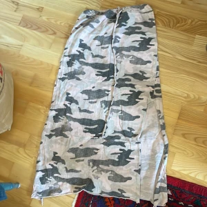 Camouflage mönstrad kjol med slits - Säljer en långkjol med grått och beige camouflage mönster. Kjolen har en slits på sidan och ett knytband i midjan för justerbar passform. Perfekt för en avslappnad stil.