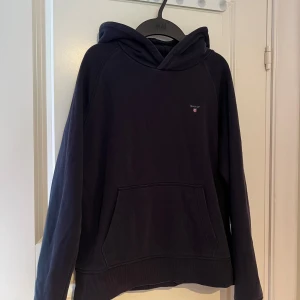 Mörkblå hoodie från Gant - Säljer en snygg mörkblå hoodie från Gant med klassisk design. Tröjan har en känguruficka framtill och en bekväm huva.