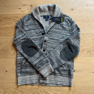 Ralph Lauren kofta i st XS - Säljer en stilren gråm kofta från Ralph Lauren. Koftan har en snygg sjalkrage och knappar framtill. Den är dekorerad med mörkare armbågslappar och har ribbade muddar. Perfekt för en avslappnad men ändå elegant look. Riktig stilren! 