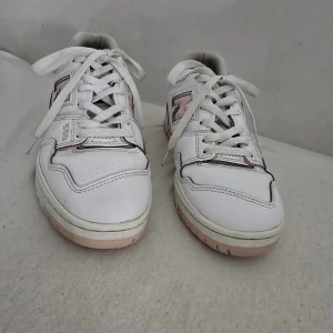 Vita och rosa sneakers från New Balance - Säljer ett par vita New Balance 550 sneakers med rosa detaljer. Skorna har snörning och en bekväm passform. Perfekta för en stilren look med en touch av färg.