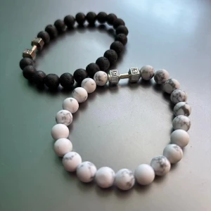 Armband - Två stilrena armband med pärlor, ett i svart och ett i vitt med marmorerat mönster. Båda har en unik detalj i form av en silverfärgad hantel. Perfekt för att ge en sportig touch till din outfit.