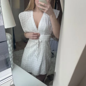 Vit kortklänning med knyt och öppen rygg - Säljer en söt vit kortklänning med v-ringning och knyt i midjan. Klänningen har en öppen rygg med en fin rosett och korta ärmar. Perfekt för sommarens alla tillfällen!