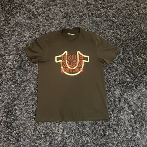 Svart t-shirt från True Religion - Säljer en svart t-shirt från True Religion med ett stort tryck i rött och orange på framsidan. T-shirten är kortärmad och har en klassisk rund hals. Perfekt för en avslappnad stil och för att ha en unik touch. Hör av er vid minsta fundering🙌