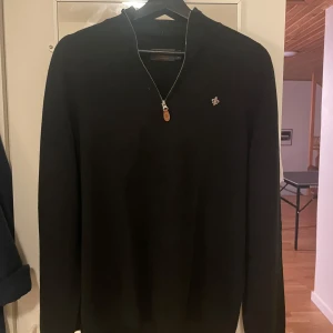 Svart Morris halfzip - Snygg svart tröja med dragkedja och en liten broderad detalj på bröstet. Perfekt för en stilren look. Tröjan har långa ärmar och en bekväm passform. Perfekt med skjorta under.