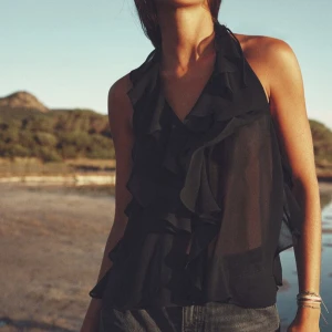 Svart ärmlös blus med volanger - Snygg svart ärmlös blus med volanger framtill. Blusen har en v-ringning och knytning i nacken, vilket ger en elegant touch. Perfekt för en stilren look. Från zara och alldrig använd 