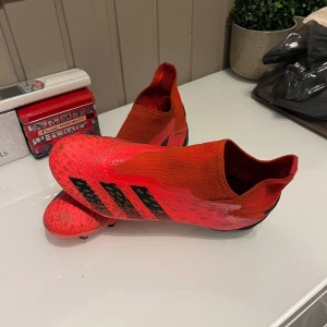 Adidas predator röda fotbollsskor  - Säljer ett par röda fotbollsskor från Adidas med svarta detaljer. Skorna har en strömlinjeformad design utan snörning och är perfekta för snabba rörelser på planen. De har en slitstark sula med dobbar för optimalt grepp. Nu pris 1500kr!