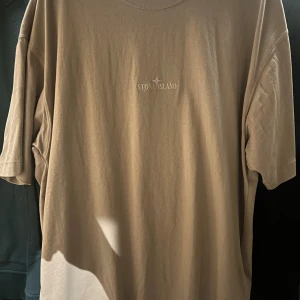 Beige t-shirt från Stone Island - Säljer en stilren beige t-shirt från Stone Island. Den har en normal passform och korta ärmar. Perfekt för en avslappnad look. Märkesloggan är diskret placerad på bröstet.