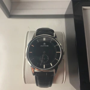 Elegant klocka från Jean Vuori - Snygg klocka från Jean Vuori med en svart urtavla och silverdetaljer. Klockan har ett svart skinnarmband och levereras i original lådan. Justerbar storlek. Nypris 2200 