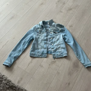 Ljusblå jeansjacka från &Denim - Snygg ljusblå jeansjacka från &Denim i klassisk modell med knappar framtill och två bröstfickor. Jackan är croppad och har långa ärmar, perfekt att slänga över en hoodie eller t-shirt. Passar dig som gillar en chill och tidlös stil.