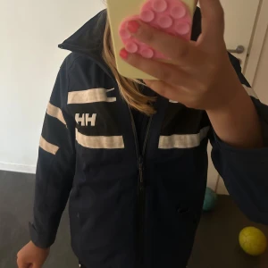 Mörkblå regnjacka Helly Hansen - Säljer en mörkblå vindjacka från Helly Hansen med vita detaljer och dragkedja framtill. Jackan har huva, långa ärmar och tydliga loggor på bröstet och ryggen. Perfekt för blåsiga dagar och snygg till streetwear-stilen. Nypris ca 2000 kr. Pris kan diskuteras!! 💕