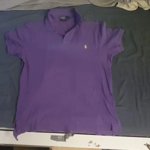 Lila pikétröja från Ralph Lauren L - Säljer en lila pikétröja från Polo Ralph Lauren i storlek L, custom fit. Klassisk piké med krage, korta ärmar och gul broderad logga på bröstet. Perfekt för dig som gillar stilren och sportig look.