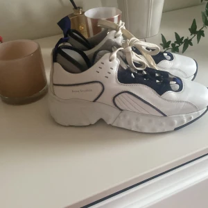 Acne Studios chunky vita sneakers - Snygga chunky sneakers från Acne Studios i vitt och marinblått skinn med coola detaljer och tjock sula. Skorna har snörning och en sportig vibe, perfekta för dig som gillar statement sneakers. Acne Studios-logga på sidan och kontrastfärgade detaljer.