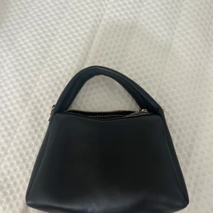 Svart handväska från H&M med dragkedja - Stilren svart handväska från H&M i syntetmaterial. Väskan har en mjuk, lite trekantig form med ett rejält handtag och guldfärgad dragkedja upptill. Snygga kontrastsömmar och enkel design som passar till många outfits.
