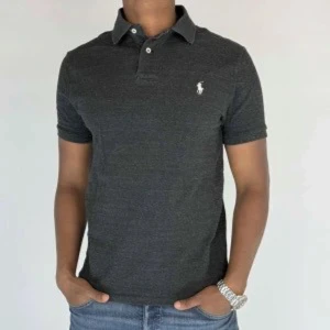 Mörkgrå pikétröja från Polo Ralph Lauren - Snygg mörkgrå pikétröja från Polo Ralph Lauren med klassisk krage och två knappar framtill. Tröjan har korta ärmar och den ikoniska broderade loggan på bröstet. Tillverkad i mjuk bomull som känns skön mot huden. Perfekt för en clean och stilren look.
