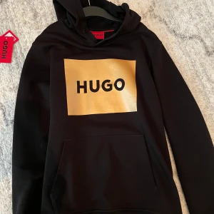 Svart hoodie från HUGO med guldtryck - Svart hoodie från HUGO med stort guldigt tryck och svart logga på bröstet. Klassisk huva och magficka framtill. Perfekt för dig som vill sticka ut med en snygg och modern streetstyle. Mjuk och skön känsla, passar till många outfits. 