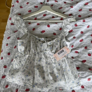 Blommig volangtopp från Nelly - Supersöt blommig topp från Nelly i ljusblått och vitt med volangärmar och spetsdetaljer. Toppen har öppen rygg med knytband och en luftig, flowy passform. Perfekt för varma dagar när du vill känna dig extra gullig.