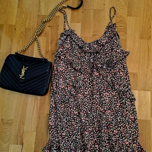 Blommig volangklänning med smala axelband - Supersöt kort blommig klänning med volanger och smala axelband. Svart botten med små blommor i rosa, vitt och grönt. Lätt och flowy modell, perfekt för varma dagar. Volangdetaljer längs byst och kjol ger en extra feminin touch.