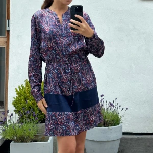 Mönstrad kortklänning med knytband - Paisleymönstrad kortklänning i marinblått, rött och vitt med lång ärm och knytband i midjan. Klänningen har en bred enfärgad marinblå bård nertill och är tillverkad i ett mjukt, lätt material som ger en avslappnad men snygg look.