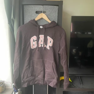 Mörkbrun GAP hoodie med broderad logga - Säljer en mörkbrun hoodie från GAP med stor broderad logga i vitt och rosa på bröstet. Tröjan har huva med snörning och en stor magficka. Perfekt för chill dagar och enkel att matcha med jeans eller mjukisbyxor.