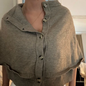 Brun/grå ponsho tröja - Snygg grå stickad kofta med stora knappar framtill och ribbade kanter. Modellen är loose och har en högre krage. Perfekt att slänga över en t-shirt eller skjorta för en chill och stilren look.