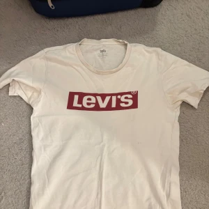 Vit Levi's t-shirt med röd logga - Vit t-shirt från Levi's med den klassiska röda loggan tryckt på bröstet. T-shirten har rund halsringning och korta ärmar. Perfekt basic-plagg med snygg streetkänsla och enkel passform.