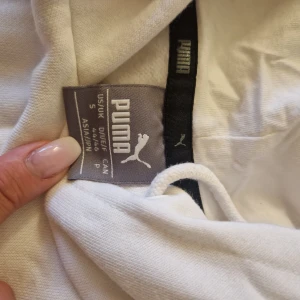 Vit hoodie från Puma med svart logga - Vit hoodie från Puma i bomull med stor svart puma-logga på framsidan. Klassisk känguruficka, huva med snörning och långa ärmar. Perfekt för en chill och sportig look.