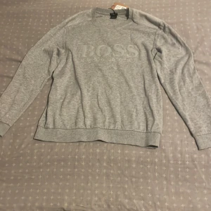 Grå sweatshirt från Hugo Boss - Snygg grå sweatshirt från Hugo Boss med stort BOSS-tryck framtill. Tröjan har rund hals, långa ärmar och ribbade muddar vid ärmslut och nederkant. Perfekt för en chill och stilren look.
