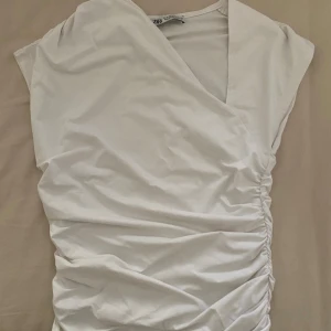 Vit omlottopp från Zara - Säljer en vit omlottopp från Zara med snygg rynkning längs ena sidan. Toppen är ärmlös och har en v-ringad design som ger en fräsch och modern look. Perfekt att styla med jeans eller kjol för en trendig outfit.