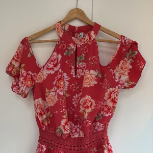 Röd blommig byxdress - Superfin röd byxdress med stora blommor i rosa och beige toner. Byxdressen har cut out-detaljer vid axlarna och korta ärmar 