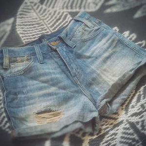 Levi's blå jeansshorts med slitningar - Snygga blå jeansshorts från Levi's med klassisk femficksdesign, slitna detaljer och uppvikta benslut. Shortsen har en cool, ljus tvätt och ett trasigt parti framtill för en avslappnad vibe. Perfekta för varma dagar och festivalhäng.