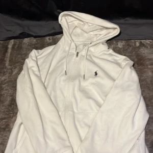 Vit hoodie från Polo Ralph Lauren - Snygg vit hoodie från Polo Ralph Lauren med dragkedja och huva. Klassisk logga broderad på bröstet och praktiska fickor framtill. Tillverkad i mjuk bomull, perfekt för en chill och stilren look. Fråga om fler bilder vid behov.