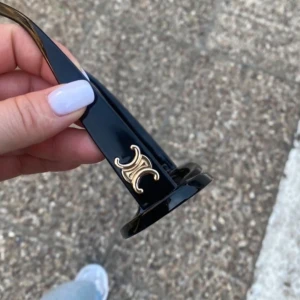 Svarta solglasögon med guldlogo - Snygga svarta solglasögon  med chunky bågar och ikoniska guldiga logodetaljer på sidorna. Glasögonen har en kantig, modern form och mörka linser som ger en cool vibe. Perfekt accessoar för att lyfta din stil.