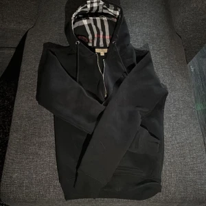 Svart Burberry hoodie - Svart hoodie från Burberry med klassiskt rutigt foder i huvan och broderad logga på bröstet. Tröjan har snörning i huvan, dragkedja framtill och långa ärmar. Perfekt för dig som gillar stilrena och exklusiva plagg. Priset inte hugget i sten! Skriv om några frågor😁😊