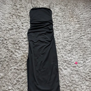 Svart långklänning med smala axelband - Säljer en svart långklänning med smala axelband och snygg twistad detalj framtill. Klänningen har en figurnära passform och är tillverkad i ett mjukt, stretchigt material som ger en clean och stilren look. Perfekt för dig som gillar minimalistisk stil.