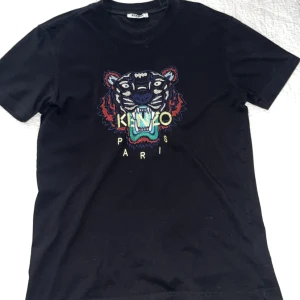 Svart Kenzo t-shirt med tigertryck - Svart t-shirt från Kenzo med ikoniskt broderat tigerhuvud och färgglad logga på bröstet. Klassisk rund hals och korta ärmar. Perfekt för dig som vill sticka ut med en statement-tee.