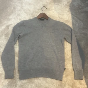 Grå v-ringad tröja från Armani Jeans - Snygg grå v-ringad tröja från Armani Jeans i mjuk ullmix. Diskret AJ-logga broderad på bröstet. Långärmad modell med ribbade muddar och klassisk passform. Perfekt för dig som gillar stilrena och enkla plagg.