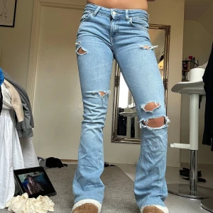 Ljusblå bootcut jeans med slitningar - Säljer ett par ljusblå jeans med bootcut-modell och snygga slitningar på låren och knäna. Jeansen har klassisk femficksdesign, hög midja och är tillverkade i stretchigt denim för en bekväm passform. Perfekta för en avslappnad och trendig look.